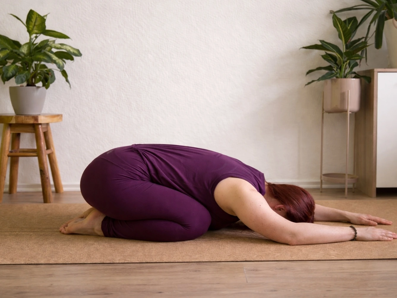 Frau in Kindhaltung (Balasana) auf einer Yogamatte, Oberkörper nach vorne abgelegt, ruhige Innenraum-Atmosphäre mit Pflanzen