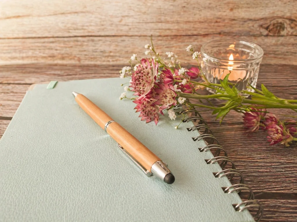 Geschlossenes Notizbuch mit Stift, Blumen und Kerze auf Holztisch – Journaling am Abend