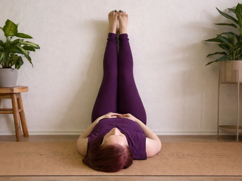 Frau in halber Kerze (Viparita Karani) mit ausgestreckten Beinen an der Wand, entspannt auf einer Yogamatte in ruhiger Umgebung mit Pflanzen