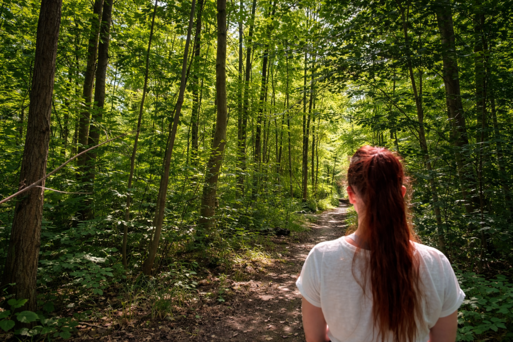 Frau auf einem Waldweg zwischen gr&uuml;nen B&auml;umen und dichtem Wald im Sonnenlicht.