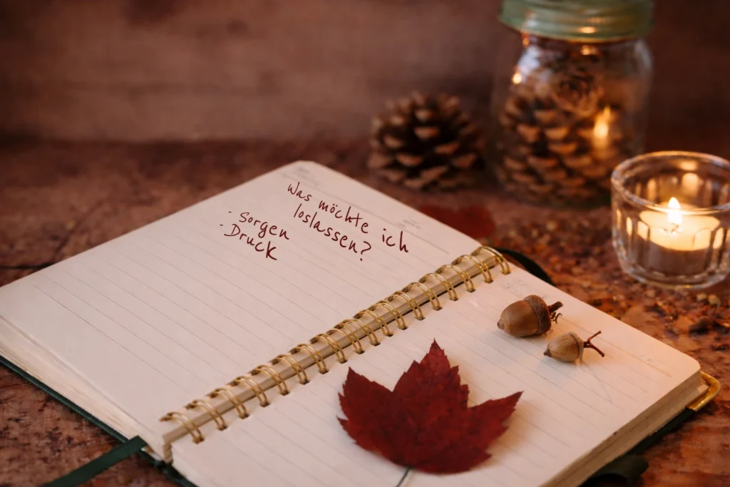 Offenes Notizbuch auf einem Holztisch mit herbstlichen Details wie Ahornblatt und Eicheln, daneben eine brennende Kerze und ein Glas mit Tannenzapfen in warmer, ruhiger Abendstimmung.