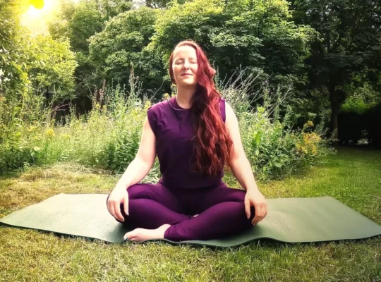 Frau sitzt nach einer Yin-Yoga-Praxis entspannt im Schneidersitz auf einer Yogamatte im Garten.