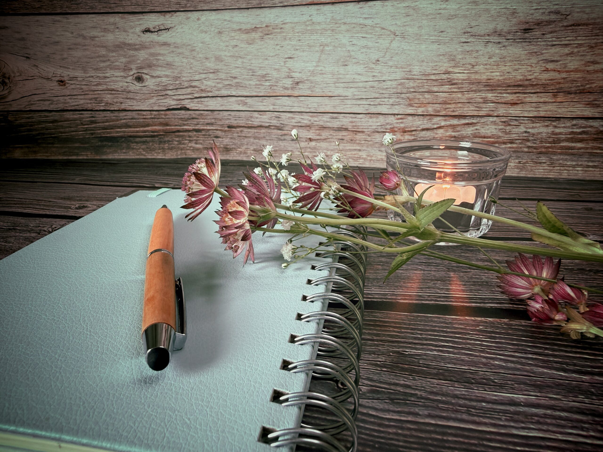 Geschlossenes Journal mit Stift, Blumen und brennender Kerze auf einem Holztisch
