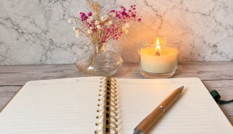 Aufgeschlagenes, leeres Journal mit einem Holzstift auf einem hellen Holztisch. Im Hintergrund stehen eine brennende Kerze und eine Glasvase mit rosa-wei&szlig;en Trockenblumen.