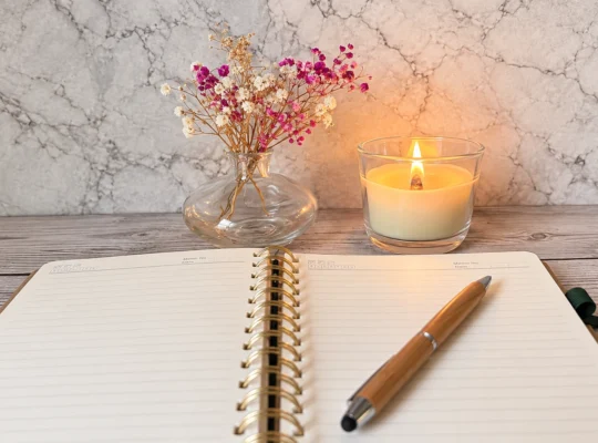 Aufgeschlagenes, leeres Journal mit einem Holzstift auf einem hellen Holztisch. Im Hintergrund stehen eine brennende Kerze und eine Glasvase mit rosa-wei&szlig;en Trockenblumen.