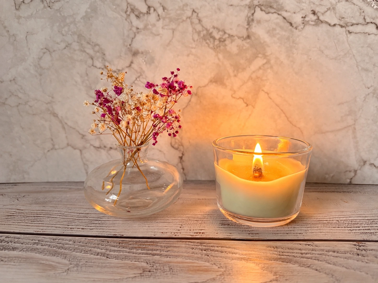Brennende Kerze im Glas neben einer kleinen Glasvase mit rosa-weißen Trockenblumen auf einem hellen Holztisch.