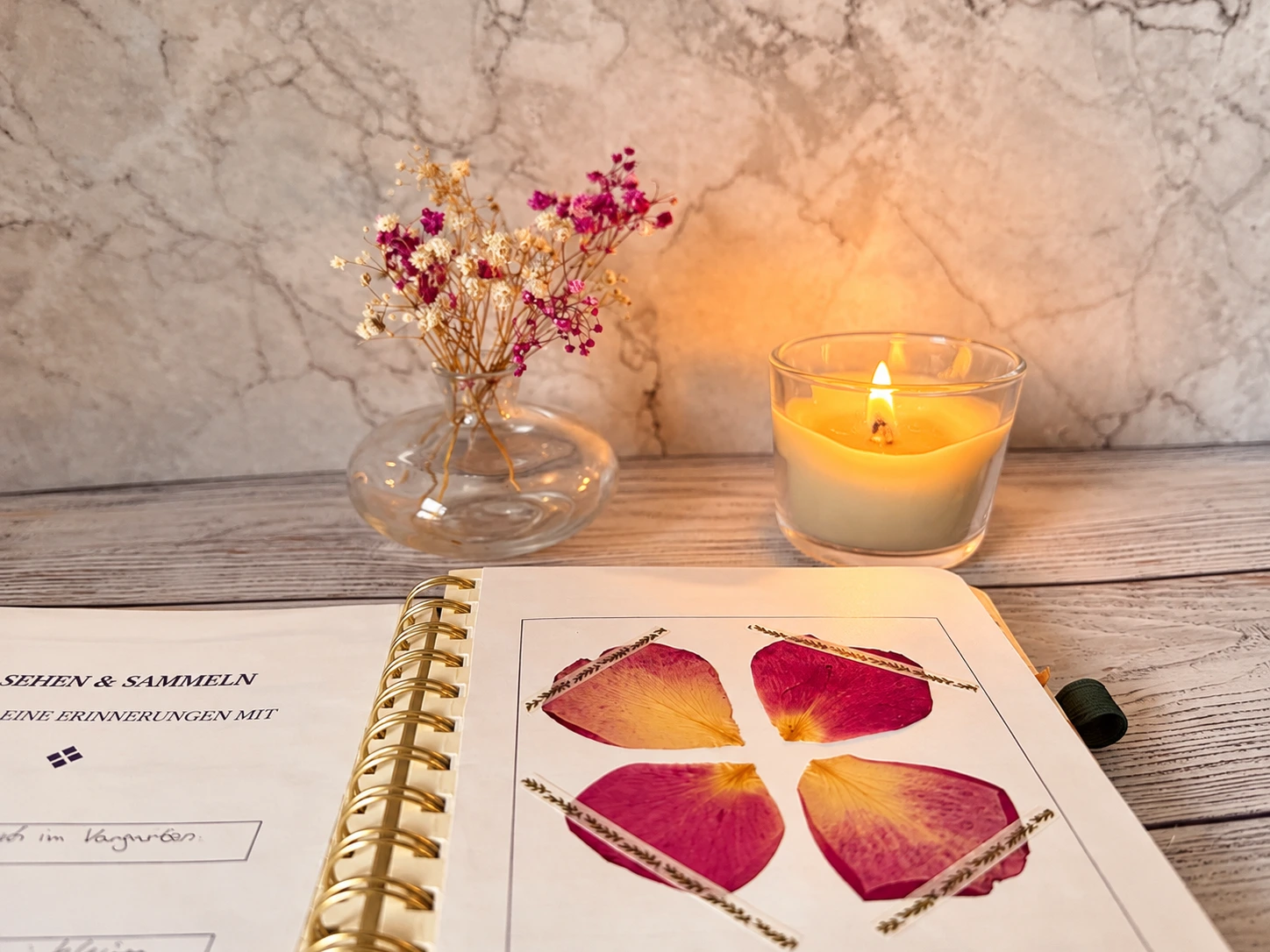Aufgeschlagenes Kreativjournal mit gepressten rosa-gelben Blütenblättern und Gräsern. Im Hintergrund stehen eine brennende Kerze und eine Vase mit Trockenblumen.