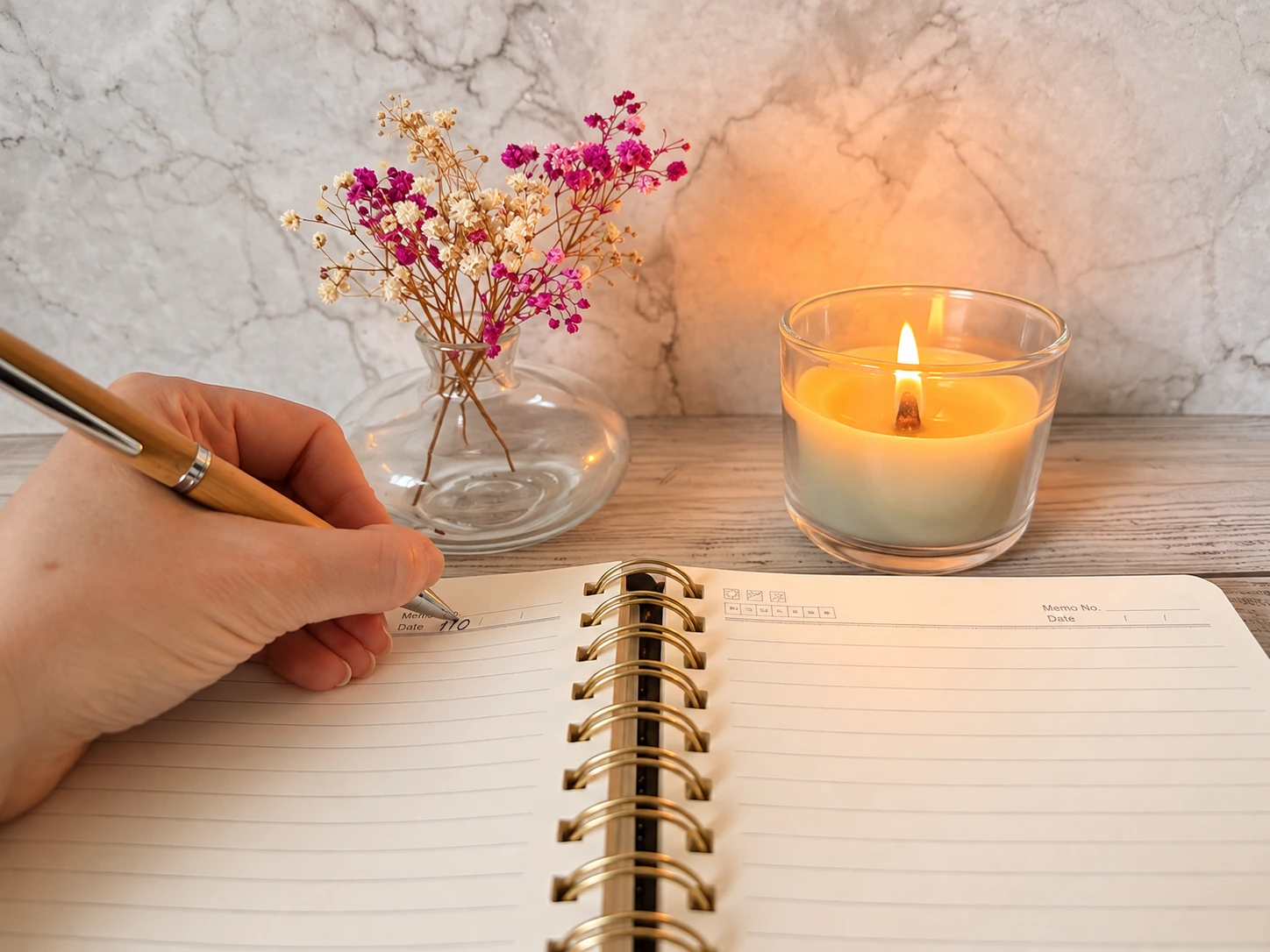 Hand schreibt mit einem Holzstift in ein aufgeschlagenes Notizbuch. Daneben stehen eine brennende Kerze und eine kleine Vase mit Trockenblumen.
